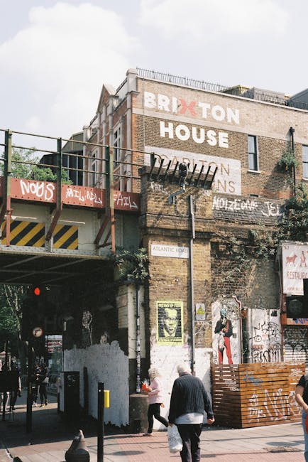 A multi-story brick building with a weathered facade featuring large, faded painted signs that spell out 'BRIXTON HOUSE' on the upper part of the wall, with the letter 'X' in red. The building has a combination of exposed brickwork and sections of worn, peeling paint. An industrial metal fire escape extends from the middle of the structure, with metal stairs and railings, partially obscured by graffiti tags on the surfaces below. Surrounding the base of the building, there is a variety of urban elements, including a small pedestrian walkway with people walking, some carrying shopping bags and engaged in conversation. To the right, a wooden barrier and a pile of discarded cardboard suggest ongoing waste clearance or rubbish collection activity. The environment appears lively, with overcast sky overhead, muted natural lighting, and a mixture of commercial and residential surroundings, consistent with areas served by private waste removal services like Rubbish Removal Brixton specializing in alternative rubbish handling methods for terrace homes or urban properties.