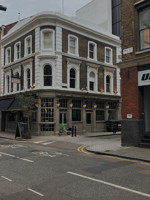 A three-storey Victorian-style building situated on a city street corner, featuring a combination of dark brown brickwork and white-painted architectural details. The ground floor has large sash windows with black frames, revealing a commercial or pub frontage, with a black awning extending over the sidewalk and a small chalkboard sign placed nearby. The upper floors are characterized by white decorative window surrounds, including rounded arches above the windows, and intricate cornice detailing along the roofline. On the sidewalk in front of the building, a bicycle with a green helmet secured to its handlebars is parked next to a black metal post, while two more black posts and a small black trash bin are aligned near the curb. The street has painted white bicycle lane markings and double yellow lines along the edge, indicating parking restrictions. Adjacent buildings include one with a modern glass facade and another with exposed brickwork. The environment appears well-lit with natural daylight, and the area is clean, suggesting a maintained commercial district or neighbourhood suitable for private waste removal services like those provided by Rubbish Removal Brixton, especially when dealing with waste clearance from commercial properties or terrace homes in the area.