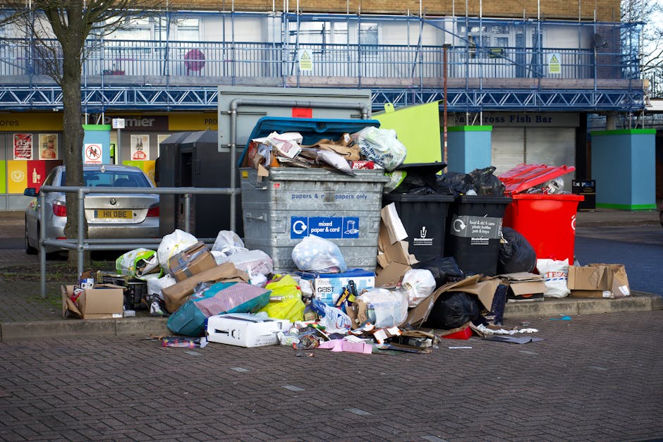 The image displays a collection of overflowing rubbish bins and scattered waste in a paved urban area, with a large grey bin partially open and filled with assorted paper and cardboard packaging. Surrounding the main bin are numerous black and red trash bags, some torn open revealing additional packaging and waste materials. Several flattened cardboard boxes and assorted paper refuse are piled on the ground nearby, alongside plastic bags containing waste. To the left, a parked silver car is partially visible behind a metal railing. In the background, a row of shopfronts with yellow and red signage is visible, along with a building covered in scaffolding and green-painted structural supports. The environment appears to be a city street or shopping area, and the scene illustrates a typical example of excess rubbish that private waste removal services, like those offered by Rubbish Removal Brixton, could manage as an alternative to on-street collection. The lighting suggests daytime with natural light illuminating the scene, emphasizing the disorderly state of the waste and the urban setting.