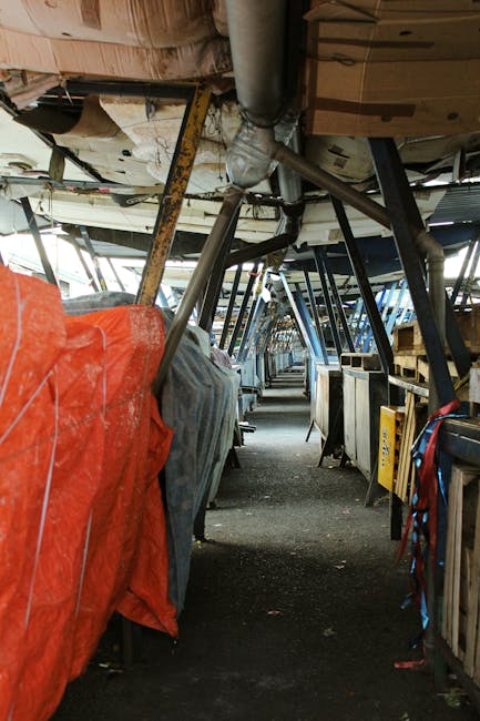 Inside the underside of a boat or large vessel, showing a narrow corridor with debris and rubbish stored along both sides. The scene includes various materials such as wooden pallets, metal pipes, and fabric covers in multiple colors, including red and gray, which appear to be protective covers or tarps. The flooring is dark and textured, possibly rubber or stone, with some dust and dirt accumulated. Structural supports and framing are visible overhead, with metal beams and hanging components that suggest an industrial or maintenance environment. The lighting is dim and natural, filtering through openings above, creating shadows and highlights on the debris and materials. This setting appears to be a storage or maintenance area, potentially involving on-site waste or rubbish management, which aligns with private or independent waste handling services provided by companies like Rubbish Removal Brixton, often dealing with large or complex debris accumulation outside conventional rubbish collection routes.