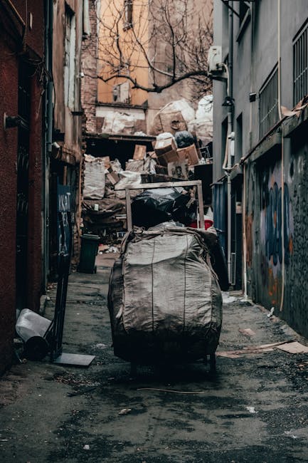 The image displays a collection of overflowing rubbish bins and scattered waste in a paved urban area, with a large grey bin partially open and filled with assorted paper and cardboard packaging. Surrounding the main bin are numerous black and red trash bags, some torn open revealing additional packaging and waste materials. Several flattened cardboard boxes and assorted paper refuse are piled on the ground nearby, alongside plastic bags containing waste. To the left, a parked silver car is partially visible behind a metal railing. In the background, a row of shopfronts with yellow and red signage is visible, along with a building covered in scaffolding and green-painted structural supports. The environment appears to be a city street or shopping area, and the scene illustrates a typical example of excess rubbish that private waste removal services, like those offered by Rubbish Removal Brixton, could manage as an alternative to on-street collection. The lighting suggests daytime with natural light illuminating the scene, emphasizing the disorderly state of the waste and the urban setting.