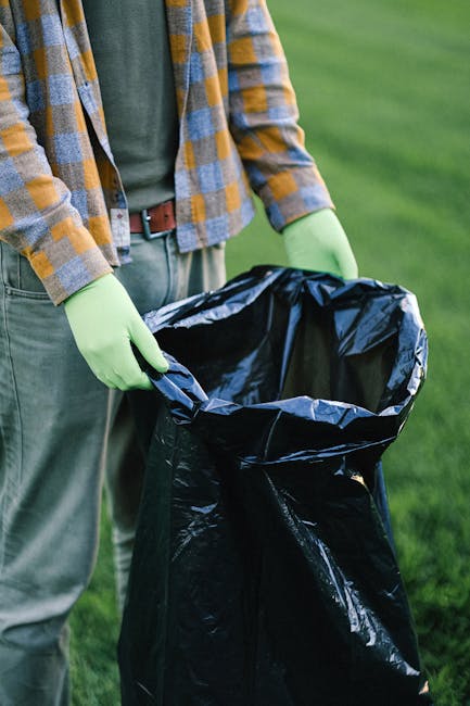 A person wearing a plaid flannel shirt in shades of yellow, blue, and white, along with green rubber gloves, is holding open a large black rubbish bag made of glossy plastic material, positioned outdoors on a paved surface surrounded by a grassy area. The person's trousers are grey with a brown leather belt visible at the waist. The scene suggests a waste collection or rubbish removal activity, with the open rubbish bag prepared for collection or disposal. The background features a blurred view of the green lawn, indicating an outdoor environment suitable for waste management activities or environmentally focused rubbish clearance services. The image aligns with the context of alternative waste handling or on-site rubbish collection, as conducted by companies like Rubbish Removal Brixton, in a neutral and professional tone suitable for accessibility and SEO focus.