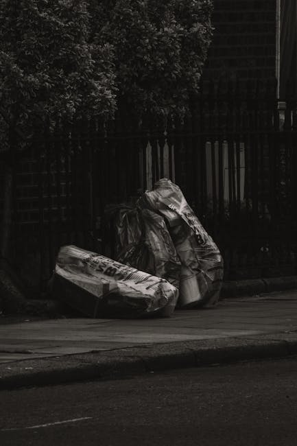 A person wearing a plaid flannel shirt in shades of yellow, blue, and white, along with green rubber gloves, is holding open a large black rubbish bag made of glossy plastic material, positioned outdoors on a paved surface surrounded by a grassy area. The person's trousers are grey with a brown leather belt visible at the waist. The scene suggests a waste collection or rubbish removal activity, with the open rubbish bag prepared for collection or disposal. The background features a blurred view of the green lawn, indicating an outdoor environment suitable for waste management activities or environmentally focused rubbish clearance services. The image aligns with the context of alternative waste handling or on-site rubbish collection, as conducted by companies like Rubbish Removal Brixton, in a neutral and professional tone suitable for accessibility and SEO focus.