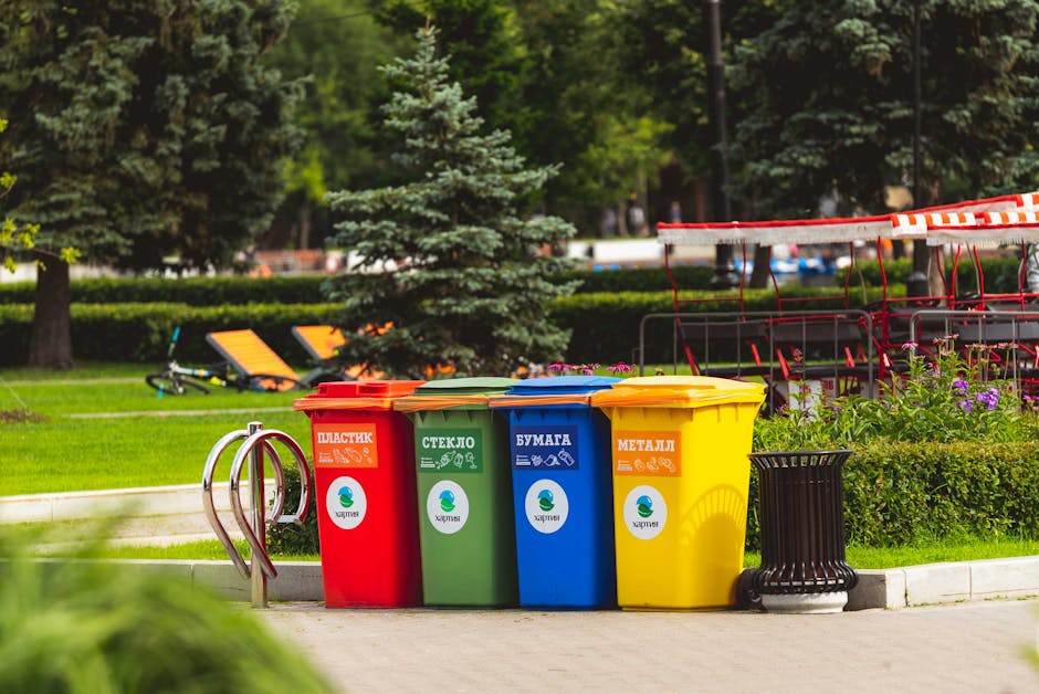 In an outdoor park setting, four colour-coded recycling bins are lined up on a paved surface. From left to right, the red bin is designated for plastics, featuring a label with the word 'ПЛАСТИК' and relevant icons; the green bin is for glass, labeled 'СТЕКЛО' with glass-related symbols; the blue bin is for paper, marked 'БУМАГА' with paper recycling icons; and the yellow bin is designated for metal waste, labeled 'МЕТАЛЛ' with metallic item icons. Each bin has a matching coloured lid and a circular white sticker with a green and blue logo at the centre. To the right of the bins, there is a small black cylindrical container with a slatted design. In the background, lush greenery with trees, shrubs, and a wooden bench with orange cushions is visible, along with a red and white striped barrier or cover, indicating a designated public space for waste sorting and recycling. The scene is brightly lit, suggesting a sunny day, emphasizing the organized waste disposal aimed at promoting environmentally responsible practices.