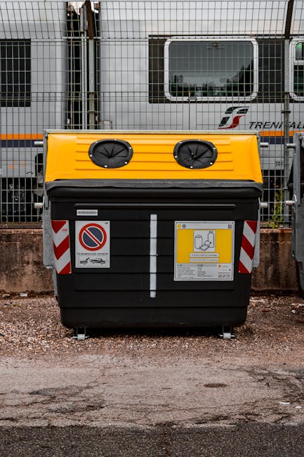 A large black waste skip with a yellow covering on the top, positioned on a gravel surface next to a concrete curb. The skip features two circular ventilation openings on the yellow lid and is fitted with reflective red and white striped warning stickers along its sides. Attached to its front is a yellow disposal instruction sign with black and white pictograms and instructions for waste disposal, as well as a no parking sign with a red border and a blue circle crossed by a red line. Behind the skip, there is a chain-link fence topped with barbed wire, revealing a background of transport containers and a grey freight train with the logo of Trenitalia, including windows and metallic surface details, suggesting an industrial or railway yard setting. The overall scene depicts an outdoor area for waste collection or rubbish removal, likely representing alternative disposal options outside of conventional municipal services, associated with private waste handling services such as those offered by Rubbish Removal Brixton.